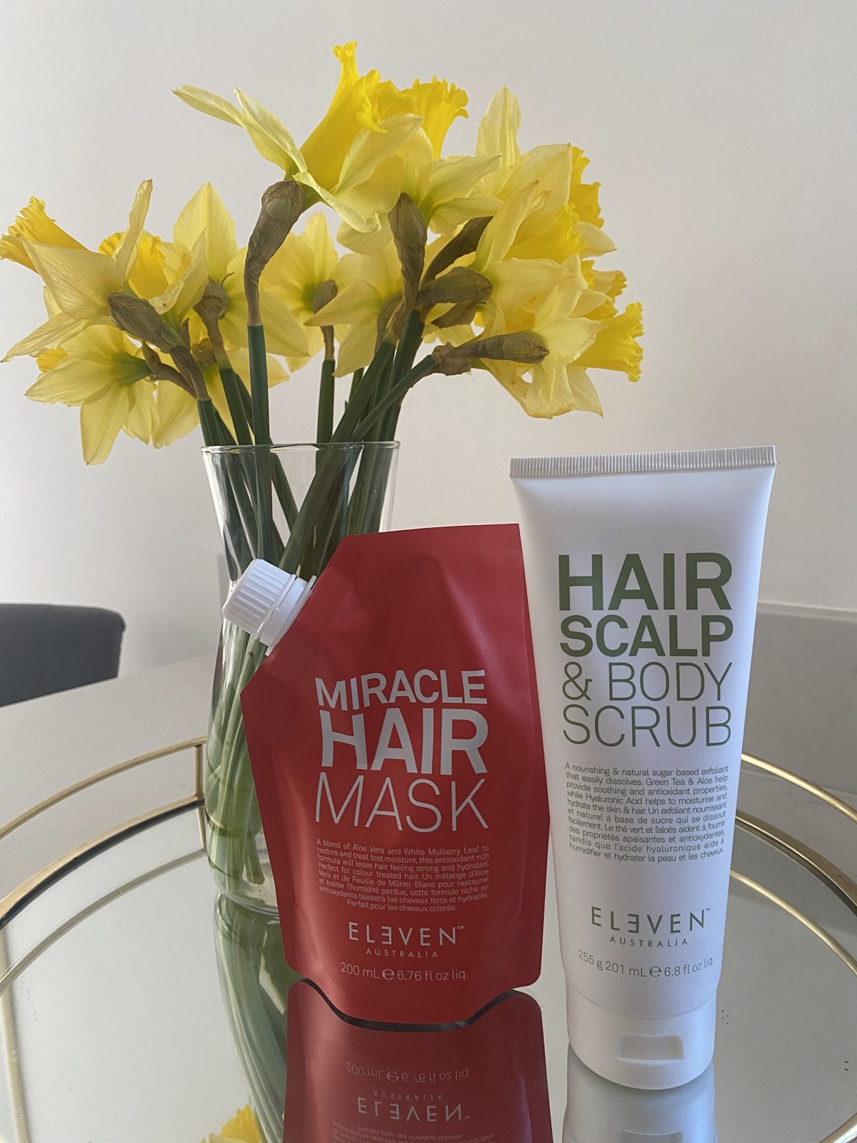 Eleven Australia Miracle Hair Mask and Hair Scalp and Body Scrub beside a vase of daffodils